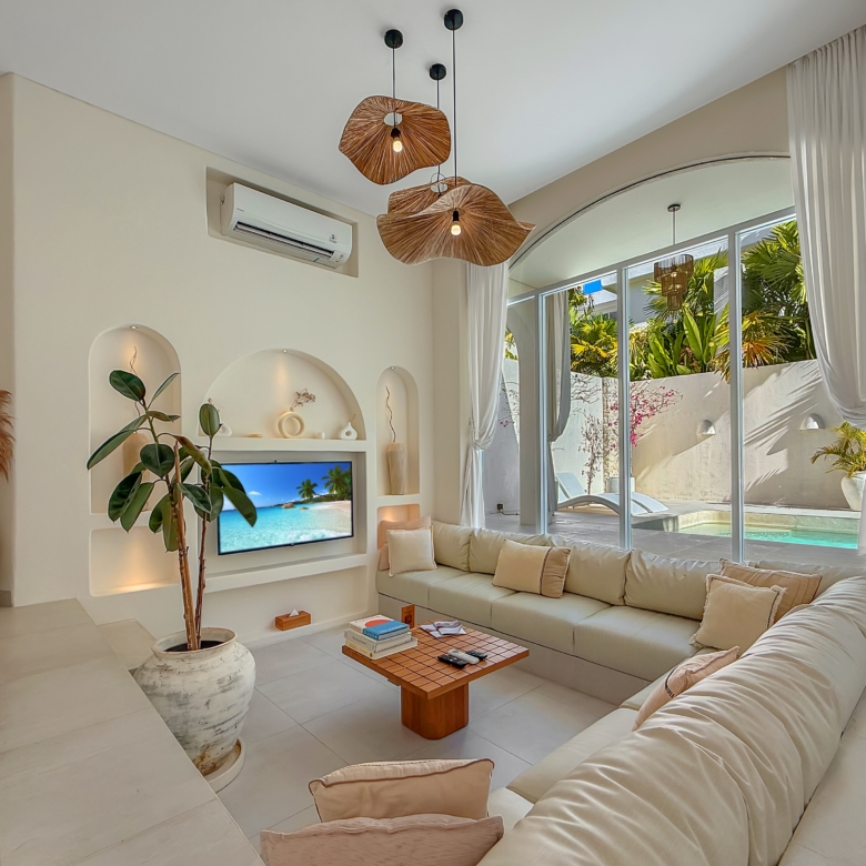 Bright living room with a white sectional sofa, hanging woven pendant lights, and large glass doors showing a pool and palm trees outside.