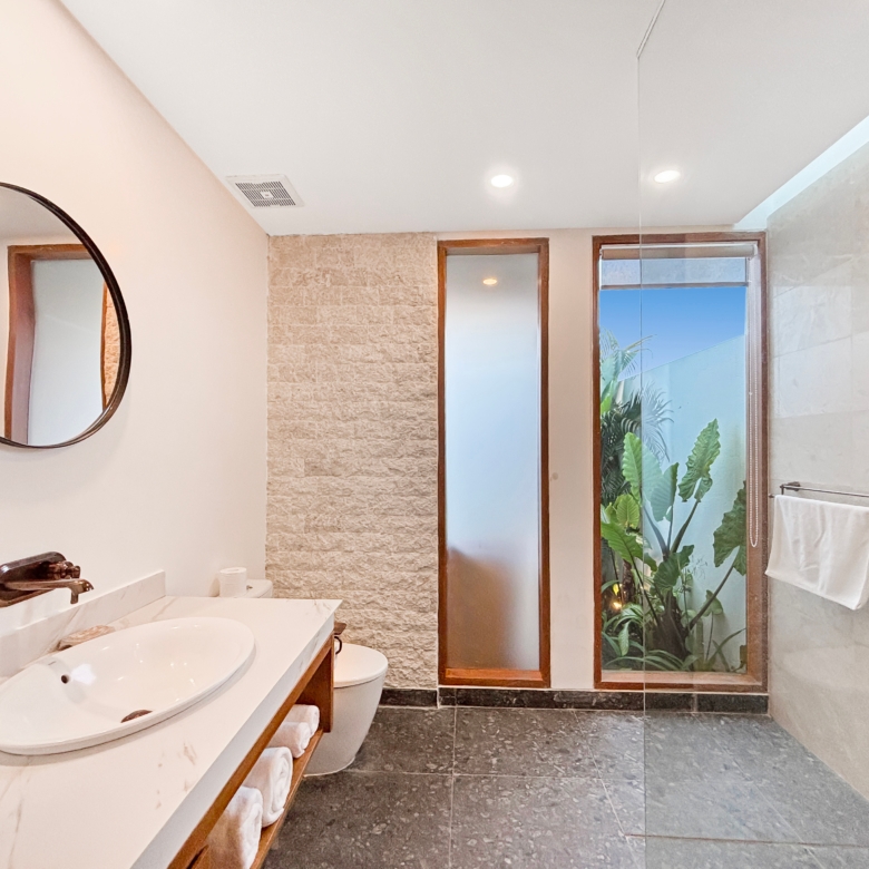 Modern bathroom with a double-sink vanity, round mirror, and glass shower door with greenery outside.