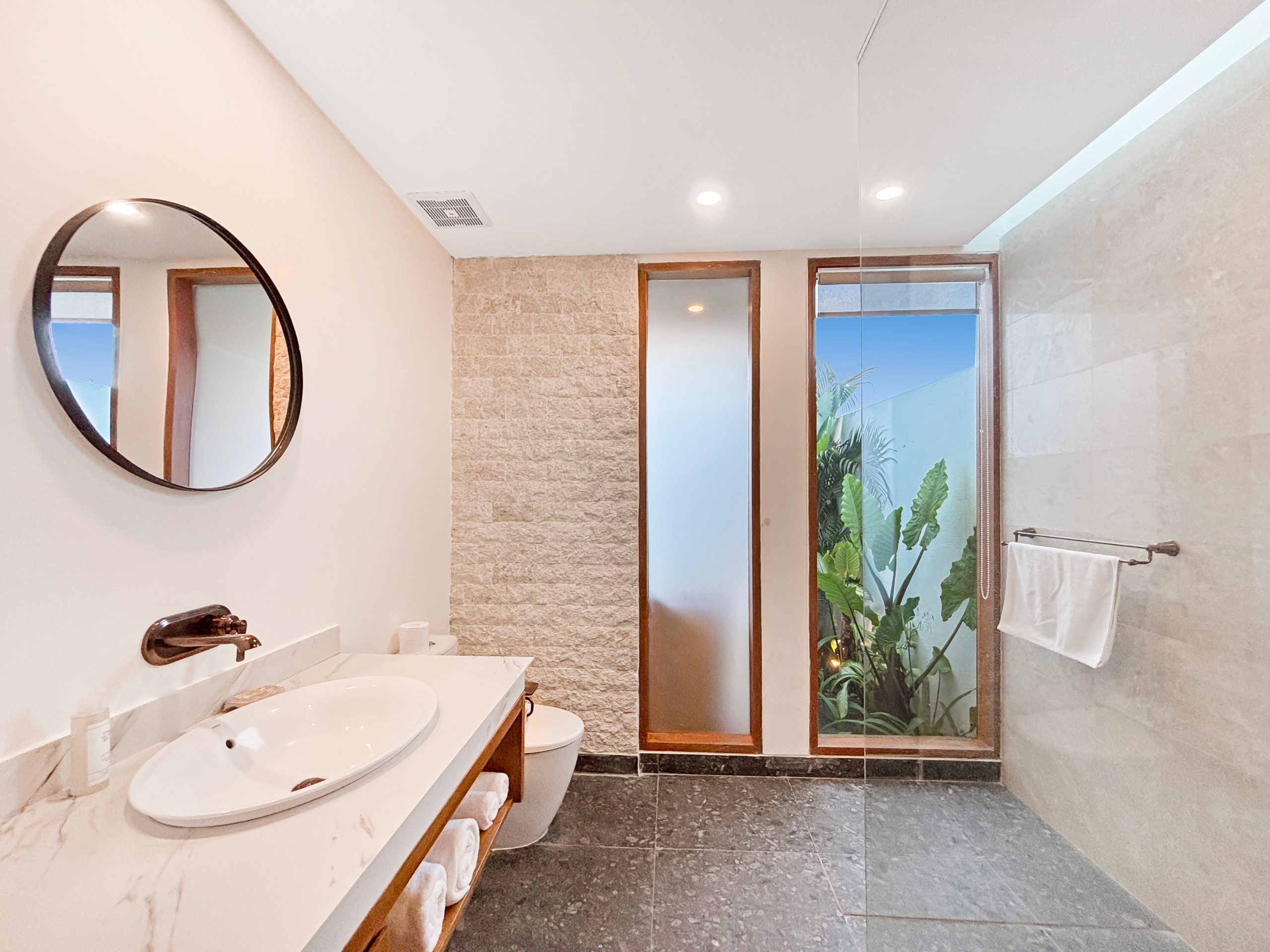 b87c43f550bacbc20c7ff44e662c64624e40ff06 Modern bathroom with a double-sink vanity, round mirror, and glass shower door with greenery outside.