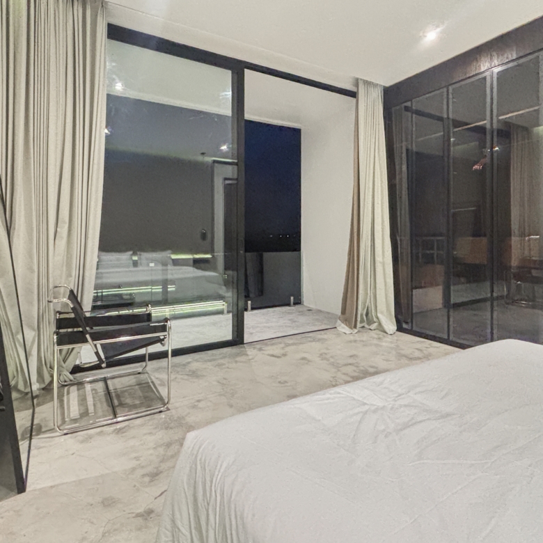 Hotel room with white bedding in the foreground, glass sliding doors, and beige curtains, showing a seating area nearby.