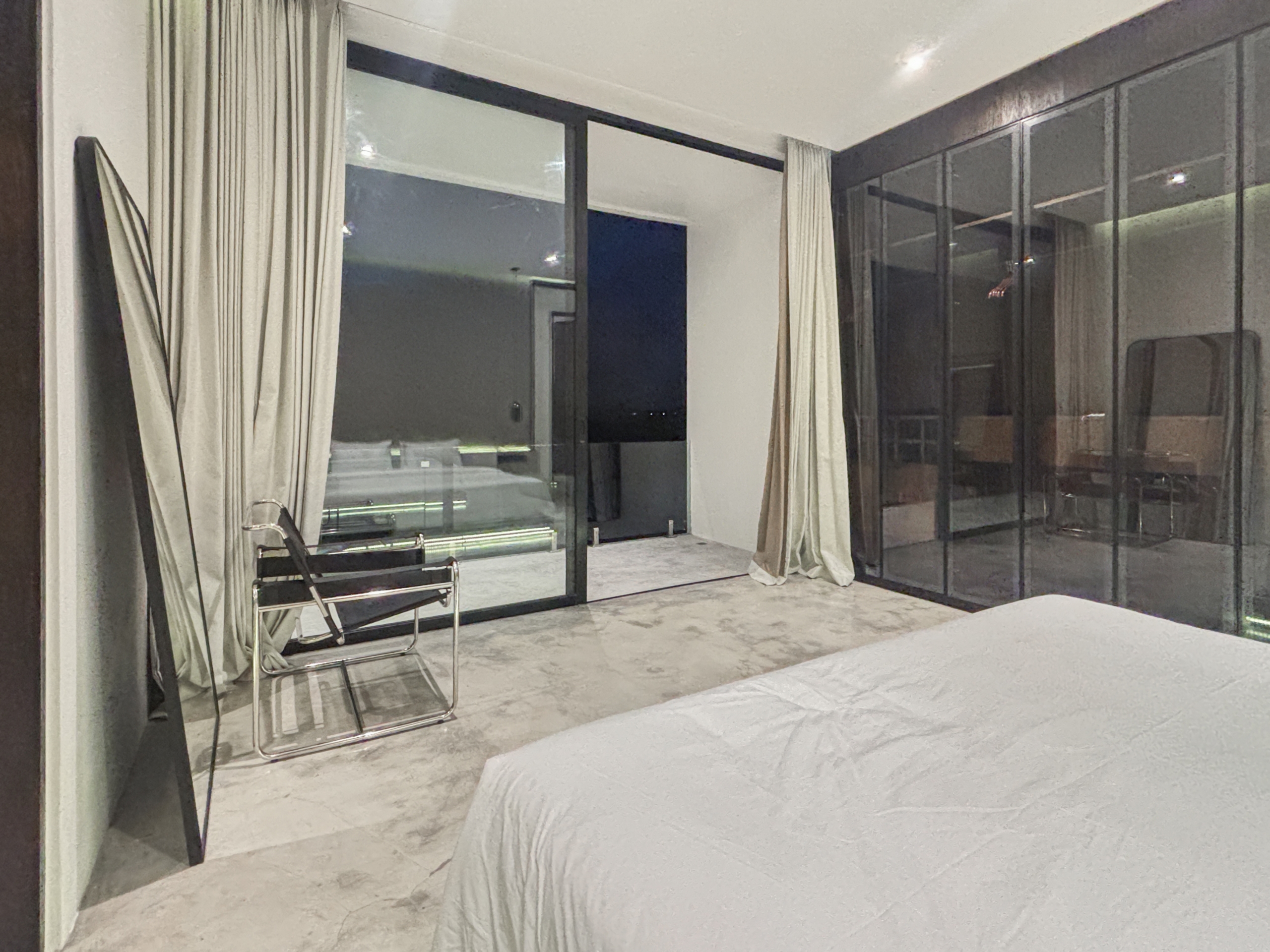 b95ca15f6749ccfa721ebfa535e74907390bb4d5_0915-6a05be0813d25 Hotel room with white bedding in the foreground, glass sliding doors, and beige curtains, showing a seating area nearby.