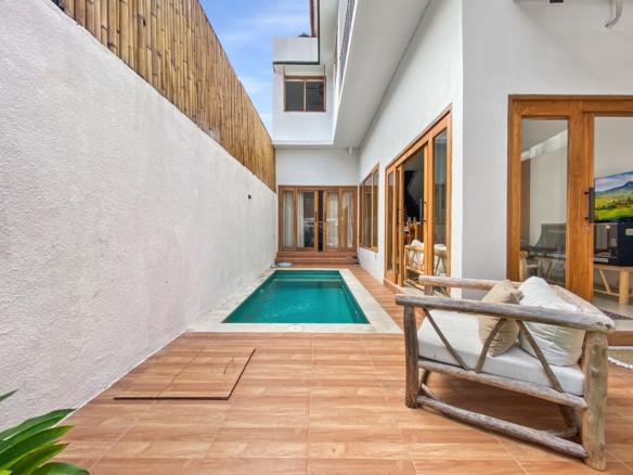 Narrow outdoor courtyard with a rectangular pool, terracotta deck tiles, white stucco walls, and wooden-framed glass doors; a wooden fence on the left and seating on the right.