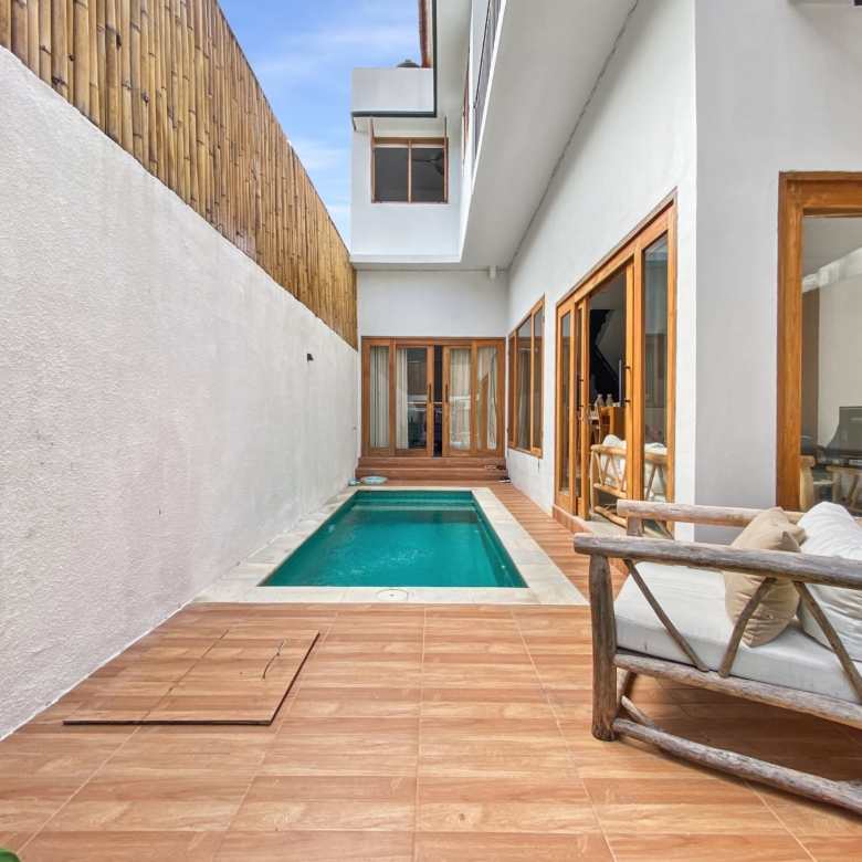Narrow outdoor courtyard with a rectangular pool, terracotta deck tiles, white stucco walls, and wooden-framed glass doors; a wooden fence on the left and seating on the right.