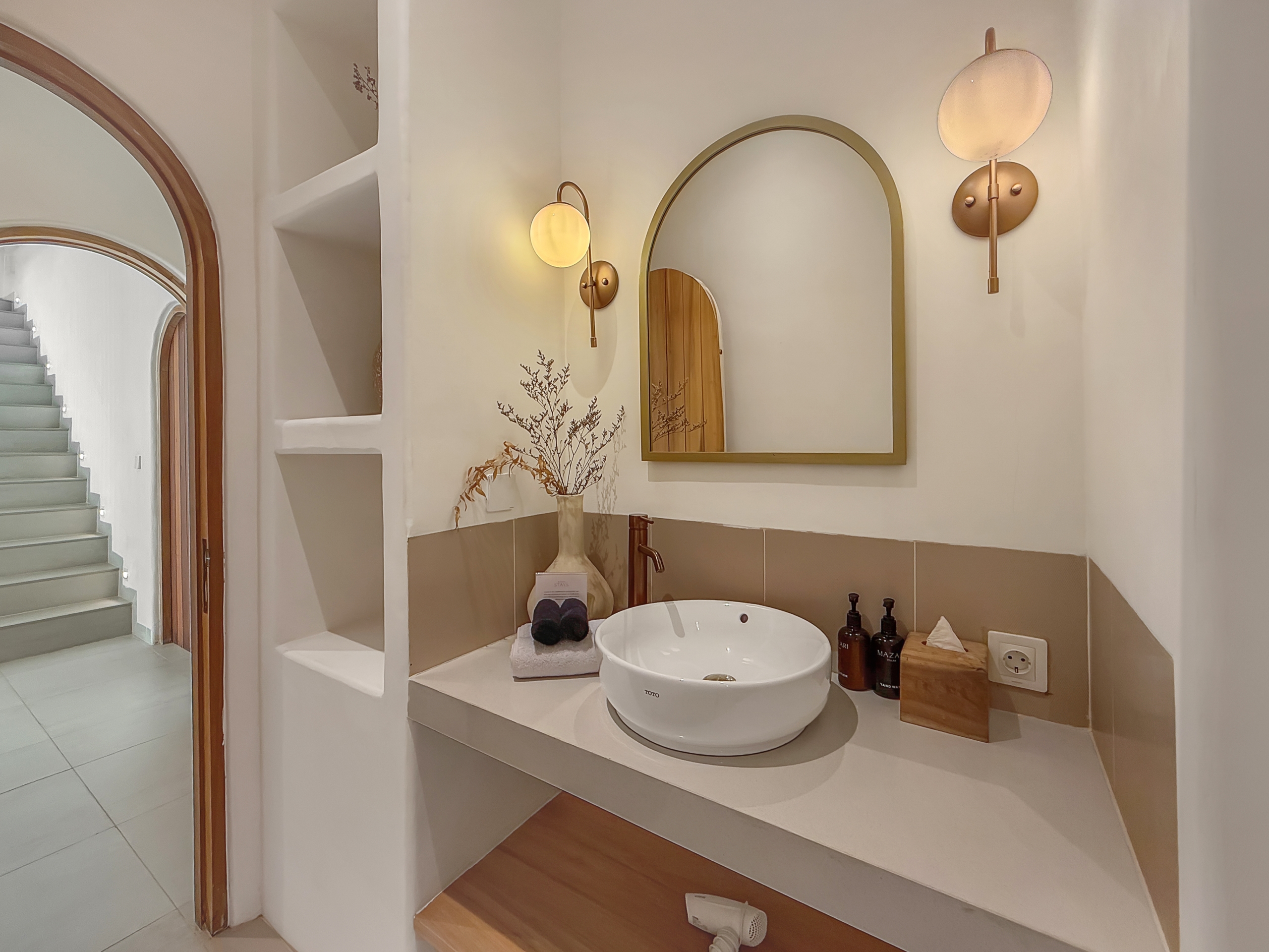 b9da435b9702b41d71c44c119da3c210b65a8100 Round vessel sink on a raised countertop with a brass-framed arch mirror and warm wall sconces in a modern bathroom vanity area.