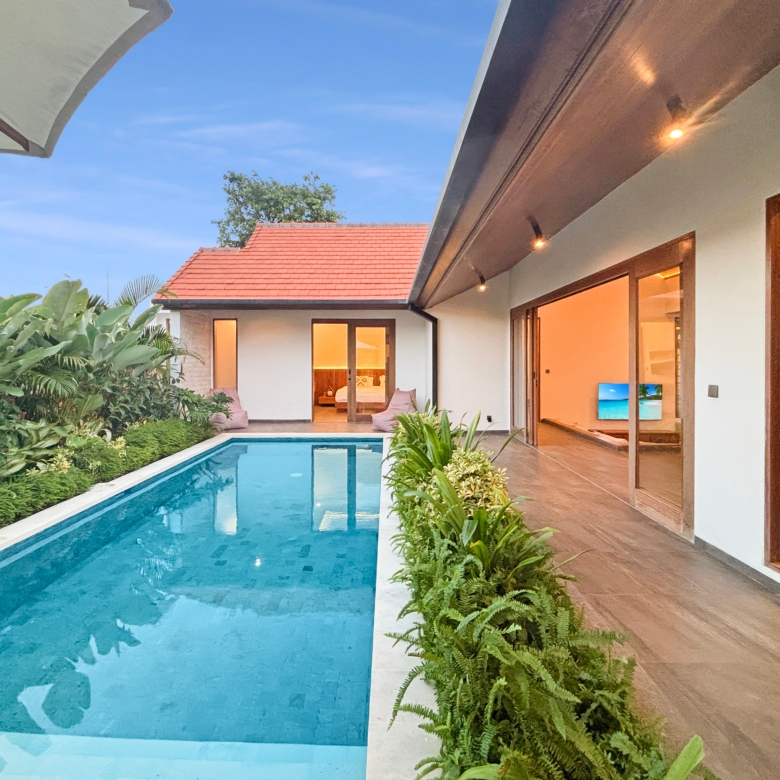 Rectangular blue pool beside a modern villa with glass doors and lush tropical plants along the edge under a clear sky.