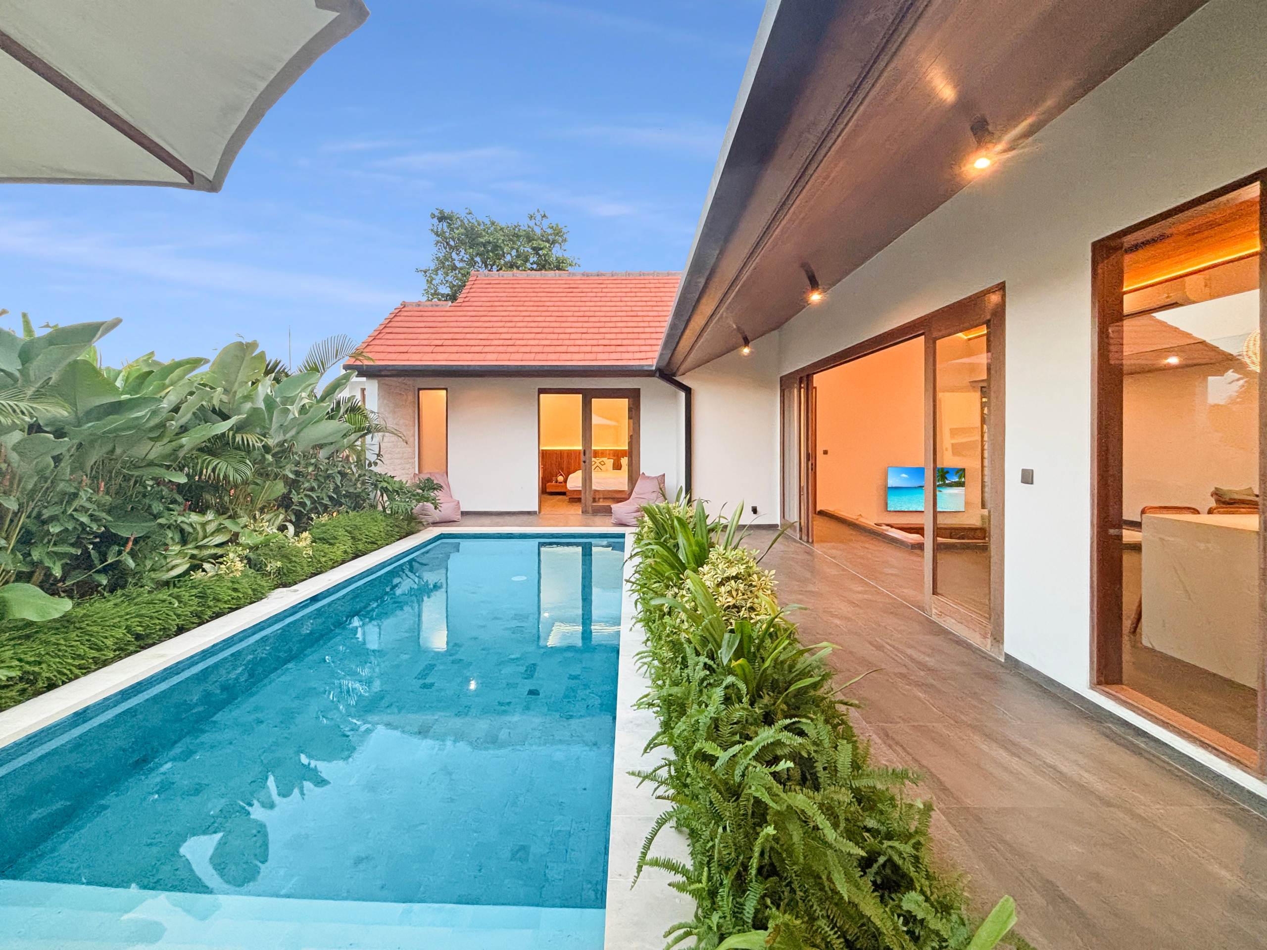 beb4900286421a90b0d619add29c83a8cb1eeb49 Rectangular blue pool beside a modern villa with glass doors and lush tropical plants along the edge under a clear sky.