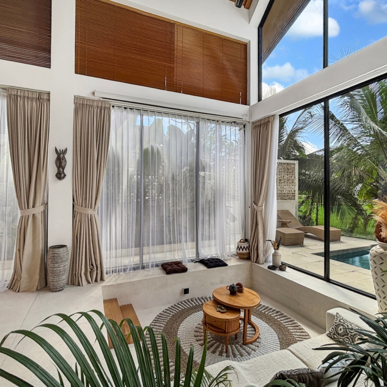 Sunlit sunroom with sheer white curtains, beige drapes, and a glass wall showing a palm-filled backyard and pool outside.