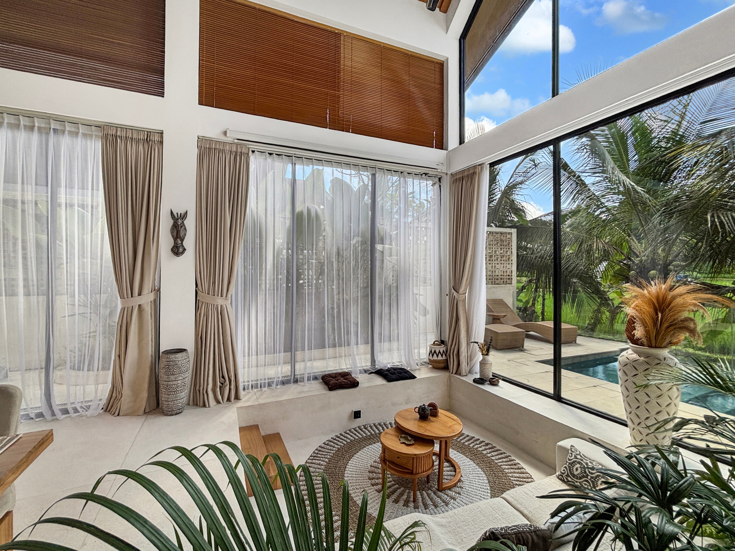 c1acfdcddffe7f281e8c0967f47e1a8331cfa293 Sunlit sunroom with sheer white curtains, beige drapes, and a glass wall showing a palm-filled backyard and pool outside.