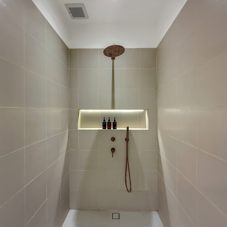 Walk-in shower with a round ceiling rain shower, tiled gray walls, and a recessed shelf holding bottles; handheld shower attached nearby.
