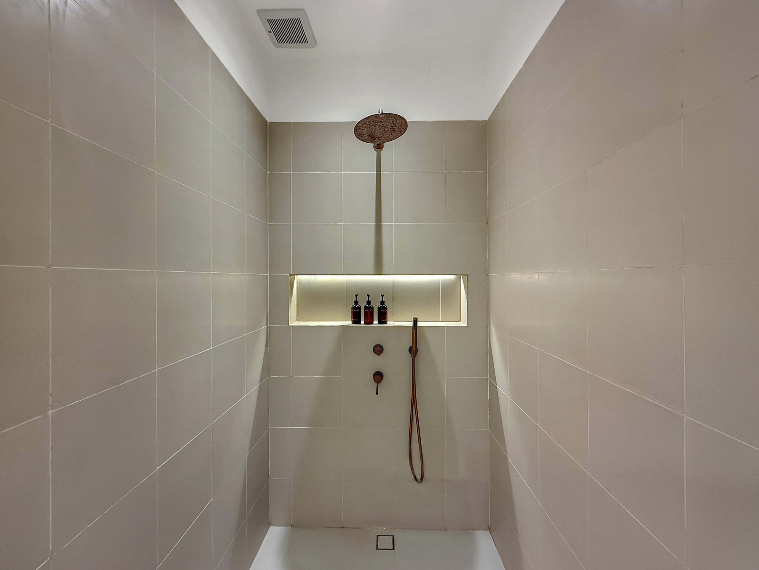 c29031a48e8f76d58810785332fa580a80ce2233 Walk-in shower with a round ceiling rain shower, tiled gray walls, and a recessed shelf holding bottles; handheld shower attached nearby.