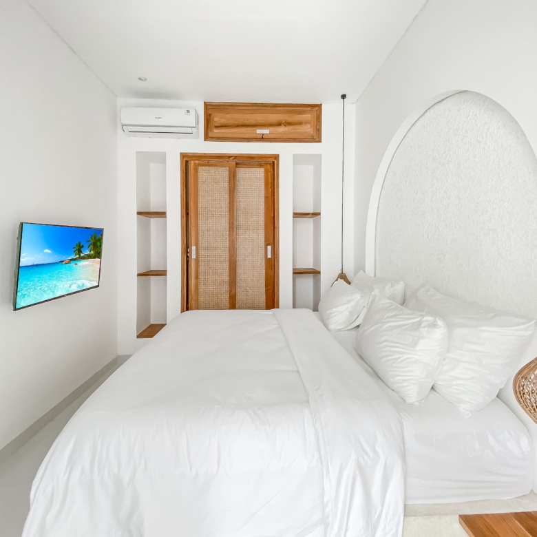 Bright white bedroom with two large white beds and wicker lamps on the right, TV mounted on the left showing a tropical beach scene, wooden door and archway in the background