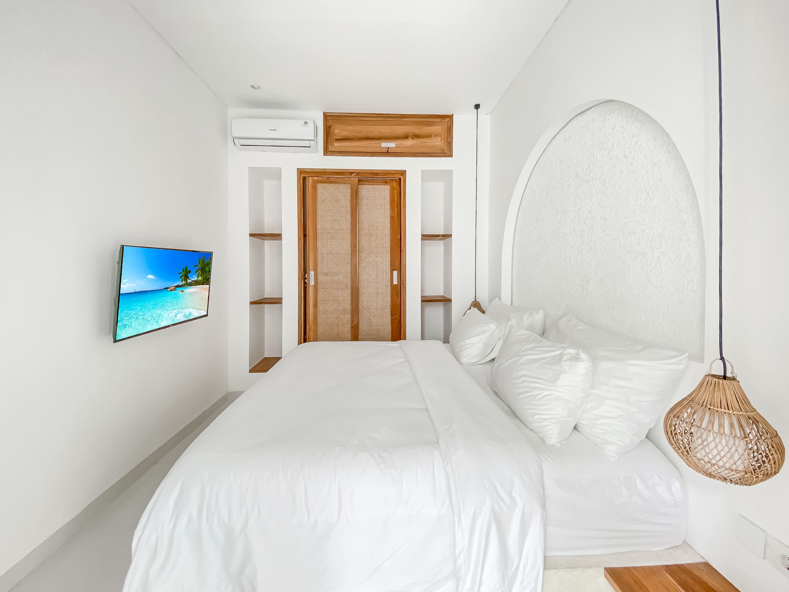 c384329df662941d11ad7b05d24a7ebba480d344 Bright white bedroom with two large white beds and wicker lamps on the right, TV mounted on the left showing a tropical beach scene, wooden door and archway in the background