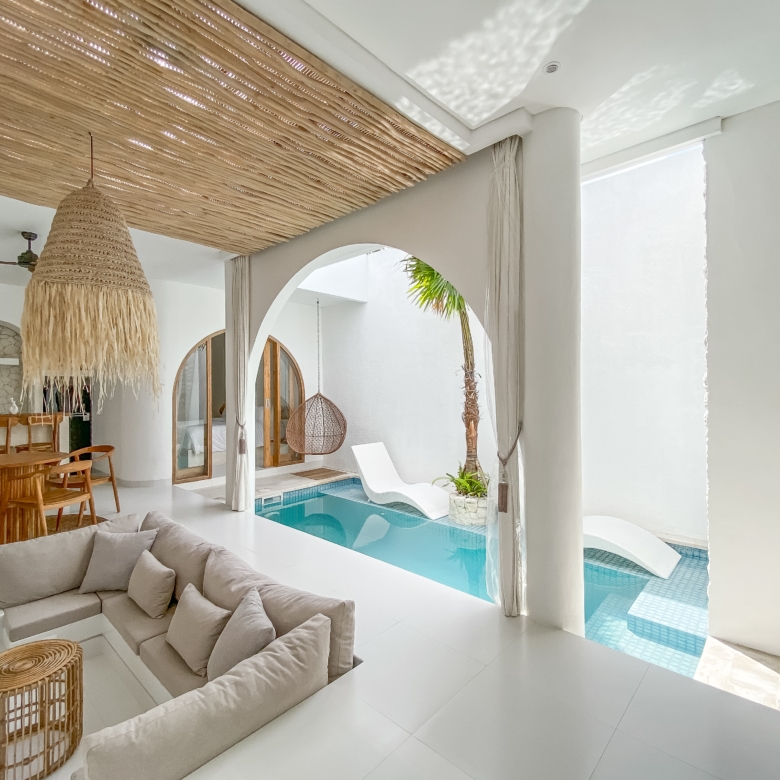 Bright open living area with a large woven pendant lamp, white walls, and a turquoise pool visible through arches.