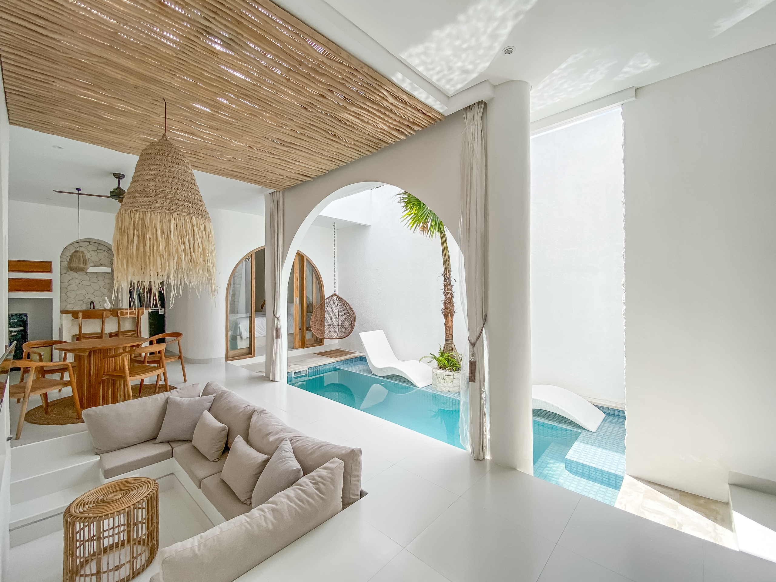 c9e2b32bed2d1ebfb48d5a54d446234b49ec4b93 Bright open living area with a large woven pendant lamp, white walls, and a turquoise pool visible through arches.