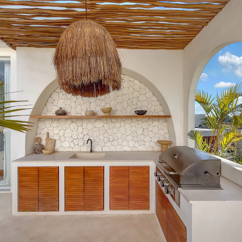 Outdoor kitchen with a built-in grill, stone backsplash, and wooden cabinet doors under a thatched hanging light.