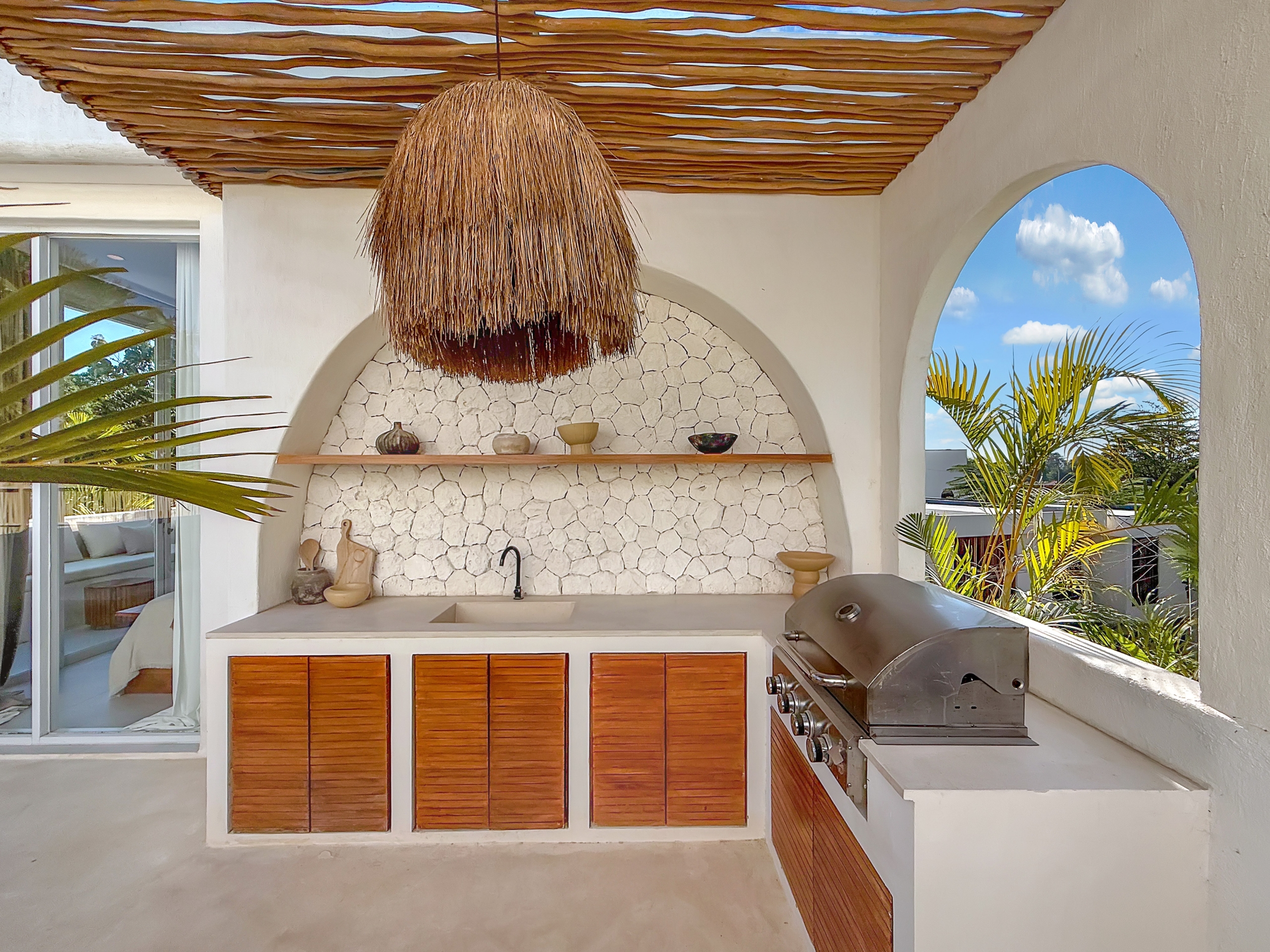 cae6a19ca03e6f00fac329560961d1694589584f Outdoor kitchen with a built-in grill, stone backsplash, and wooden cabinet doors under a thatched hanging light.