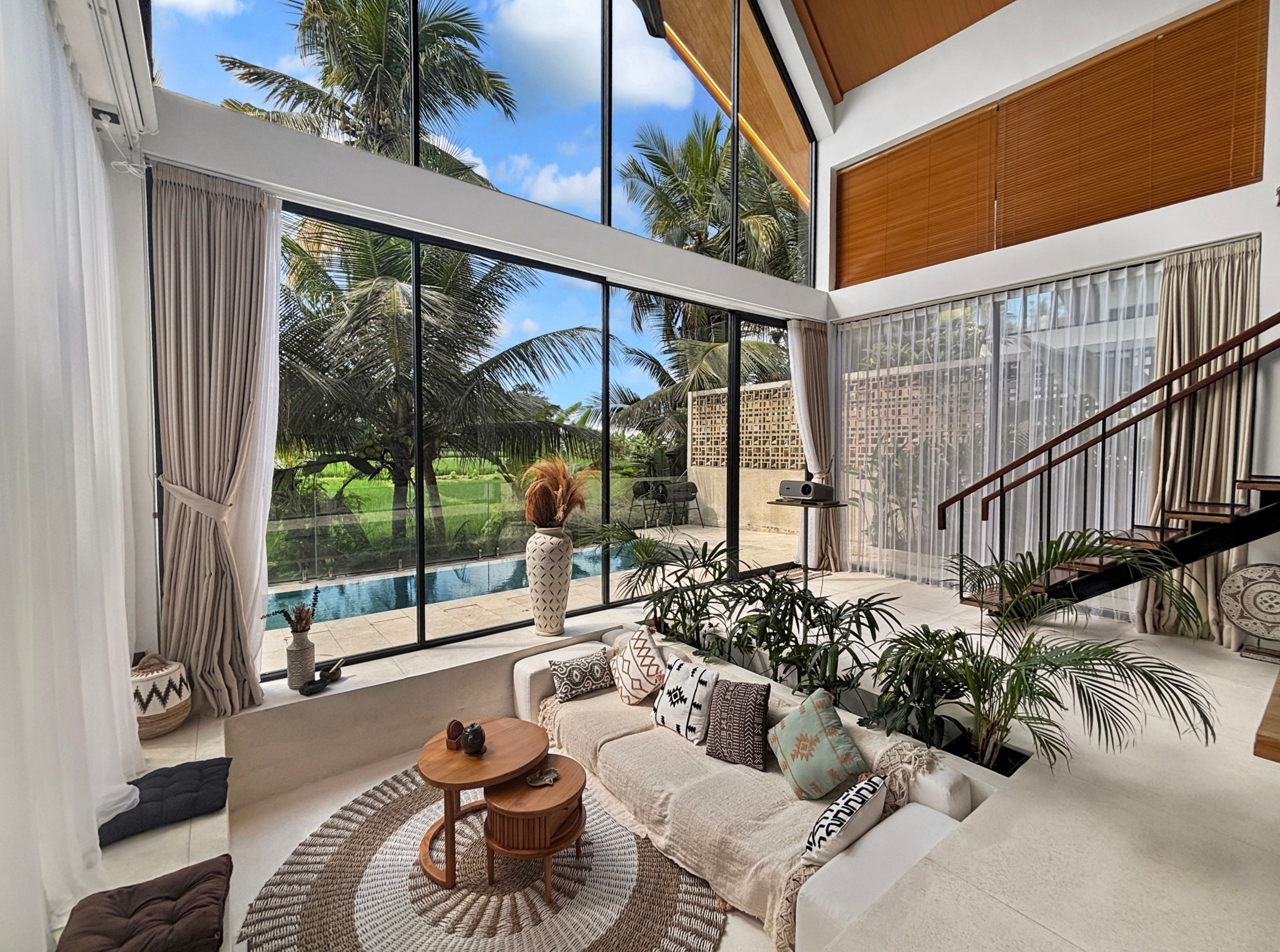 cafd48588184073c37dcbf0a42aa777f651e2e19 Bright, airy living room with floor-to-ceiling glass walls showing palm trees and a pool outside; neutral seating and woven rug.