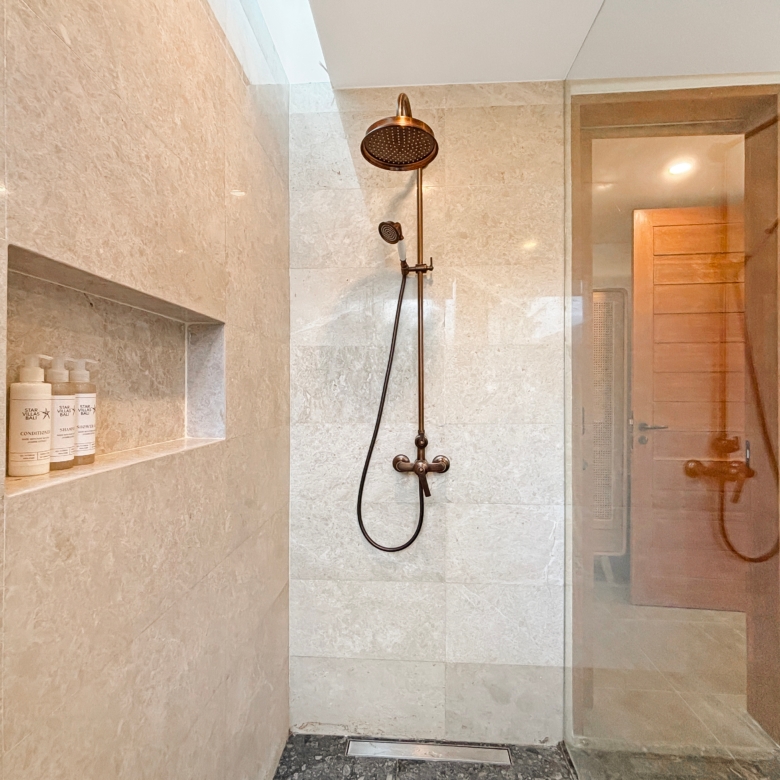 Shower area with a bronze rainfall showerhead and a handheld wand on a vertical bar, beige tiled walls, and a built-in shelf with shampoo bottles.