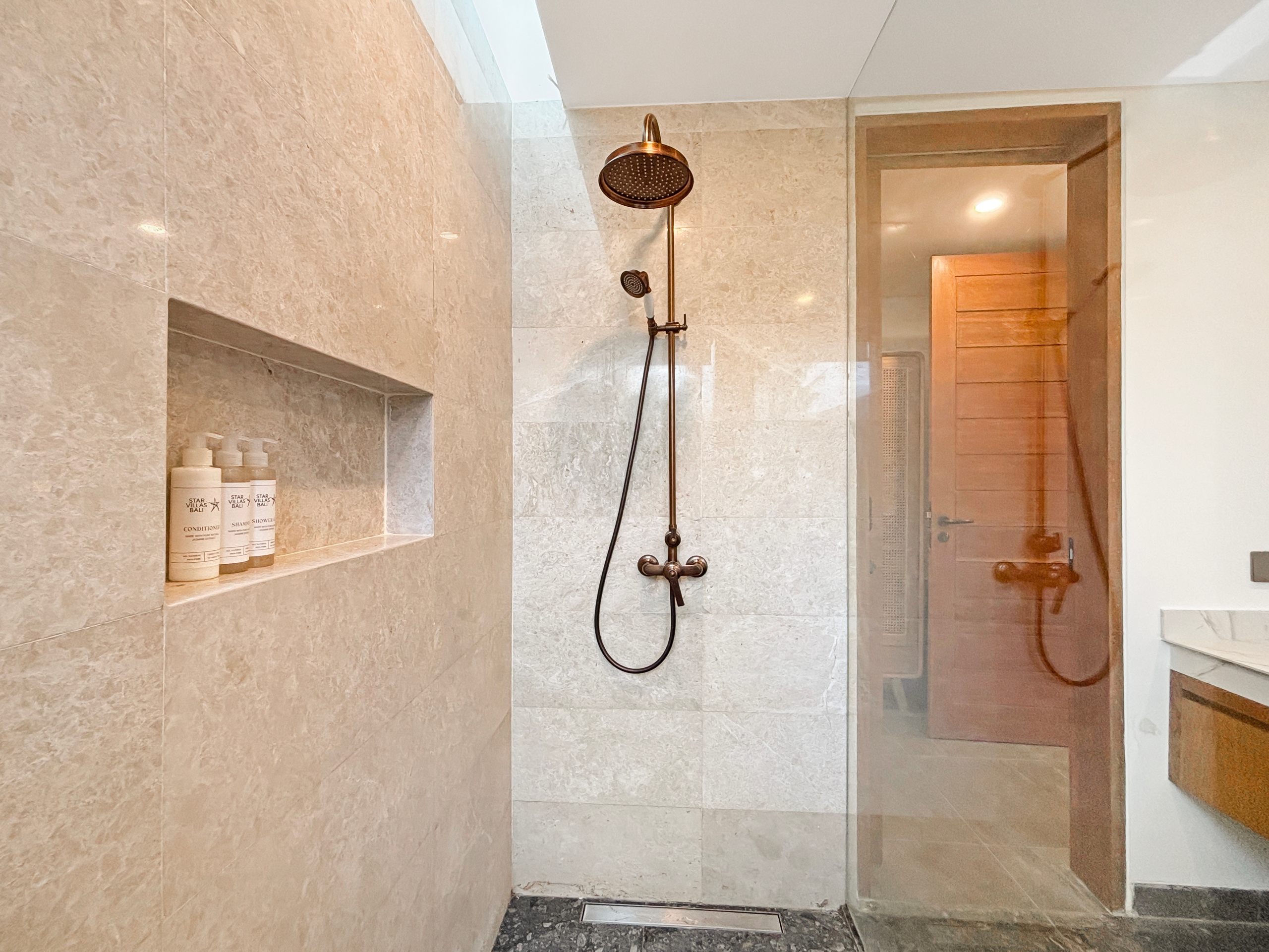 ce350fd62a7e49828675b7176703adf3976fb91a Shower area with a bronze rainfall showerhead and a handheld wand on a vertical bar, beige tiled walls, and a built-in shelf with shampoo bottles.