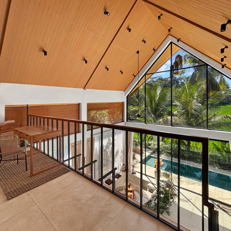 Interior loft with an orange wooden sloped ceiling and a large glass wall showing palm trees and a pool outside, plus wicker chairs and wooden furniture nearby.