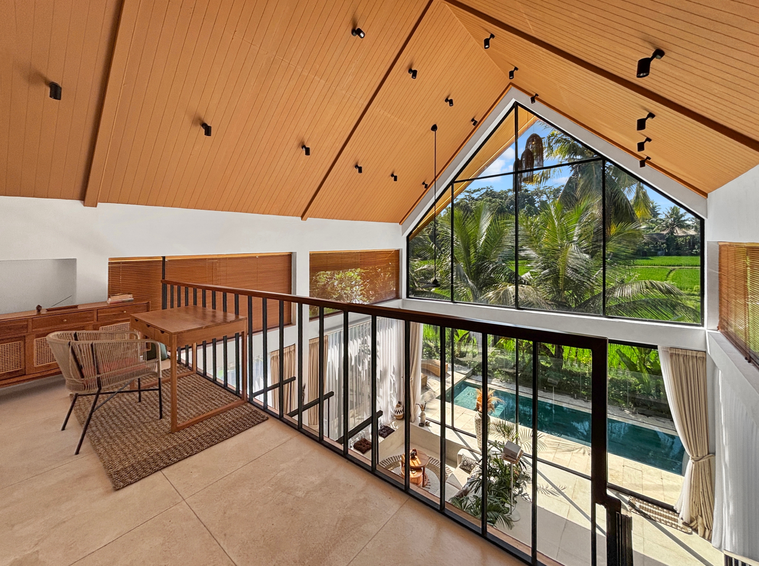 d003617981c2aa408a8f6272a5f3d77cec1f953d Interior loft with an orange wooden sloped ceiling and a large glass wall showing palm trees and a pool outside, plus wicker chairs and wooden furniture nearby.