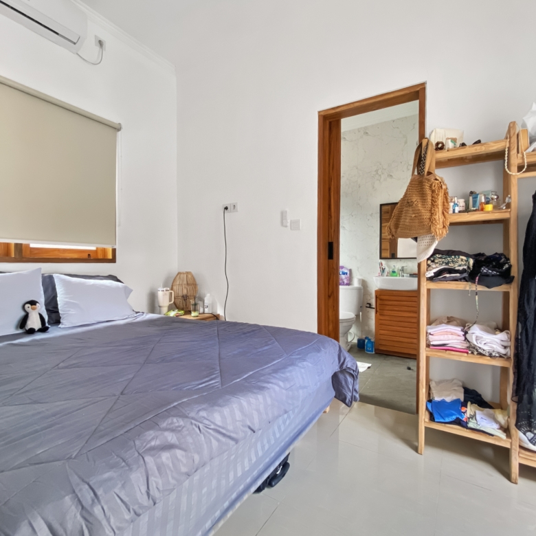 A tidy bedroom with two blue-gray beds, a small penguin plush on the left bed, and a wooden open wardrobe filled with clothes on the right side.