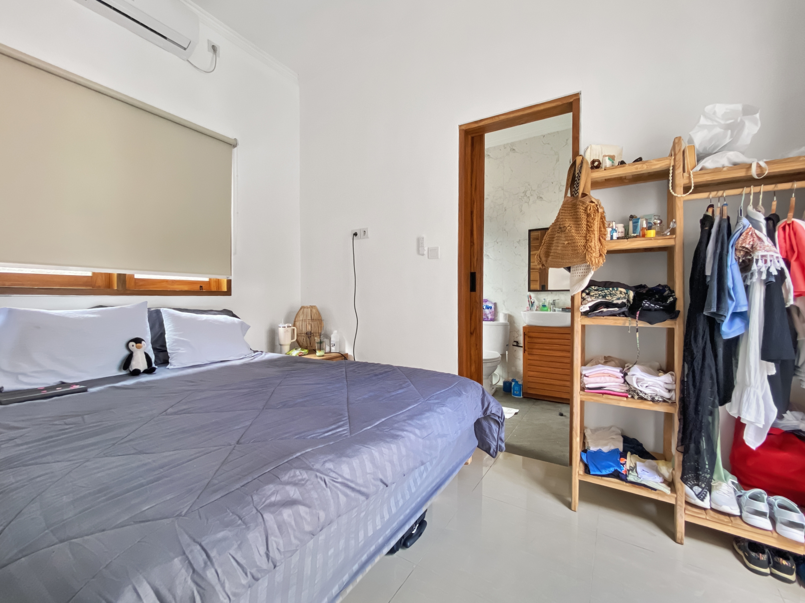 d411830160b4fd22f73d26e3478f1cda78c4b43f A tidy bedroom with two blue-gray beds, a small penguin plush on the left bed, and a wooden open wardrobe filled with clothes on the right side.