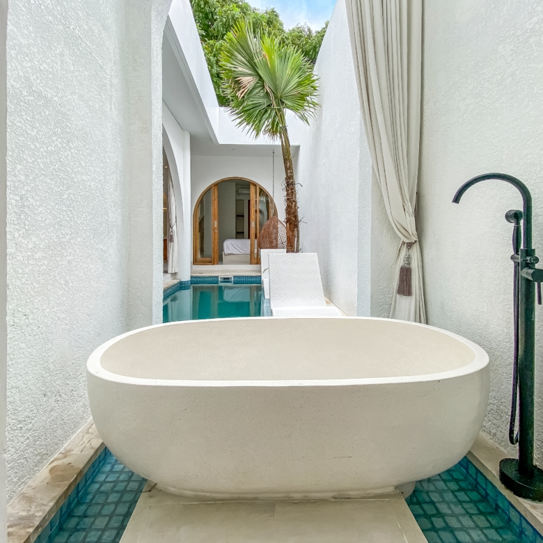 Broad view of a modern white courtyard with a large oval freestanding tub near a turquoise tiled pool, palm tree, and sheer white curtains framing an arched doorway to an indoor space.