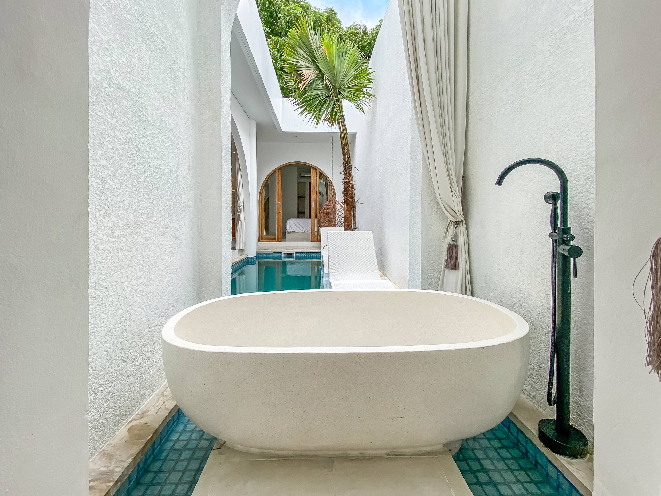 d54cbd3e5bba7017e041b6fc6edca22c0ac30d34 Broad view of a modern white courtyard with a large oval freestanding tub near a turquoise tiled pool, palm tree, and sheer white curtains framing an arched doorway to an indoor space.