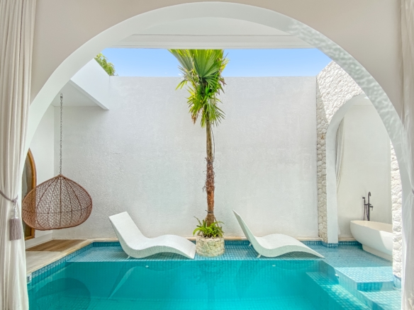 White-washed courtyard with archways, a tall palm tree, white lounge chairs, and a turquoise pool nearby.