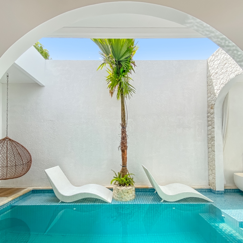 White-washed courtyard with archways, a tall palm tree, white lounge chairs, and a turquoise pool nearby.