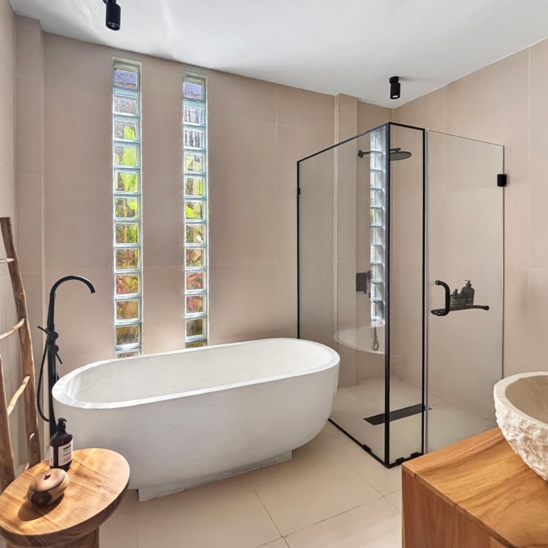 Bright bathroom with a white freestanding tub, glass‑shower enclosure, glass block windows, and a wooden ladder prop against pale walls.