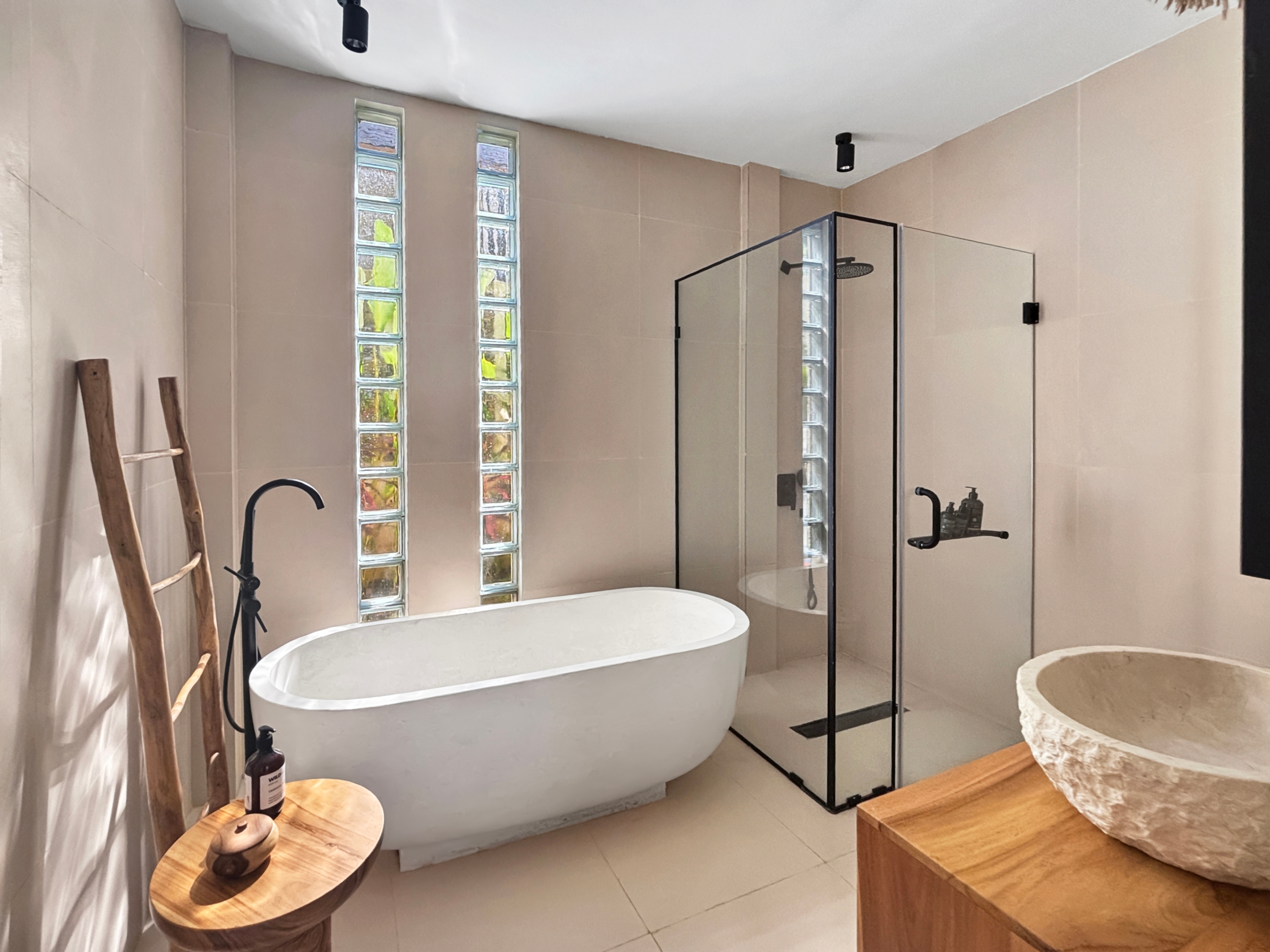 d76bacc3015921fc0e62fe75e2b2a93f72eb6c3e Bright bathroom with a white freestanding tub, glass‑shower enclosure, glass block windows, and a wooden ladder prop against pale walls.