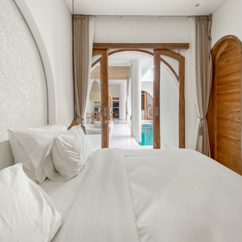 White bed with fluffy pillows in the foreground, leading to a bright hallway with wooden arches and curtains, pool visible at the end.