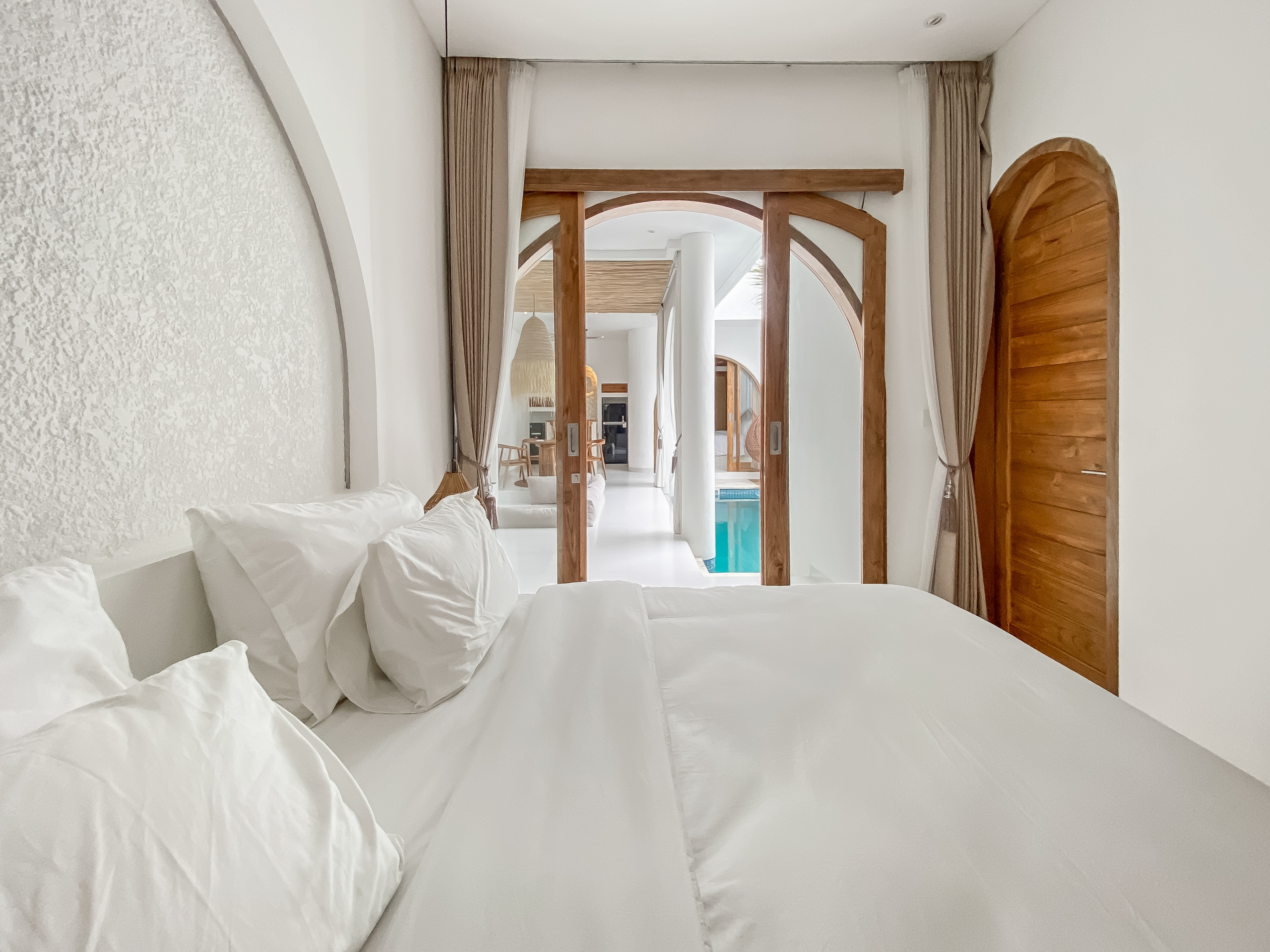 da600d27959d134d367104af75a90d2527bd4a5e White bed with fluffy pillows in the foreground, leading to a bright hallway with wooden arches and curtains, pool visible at the end.