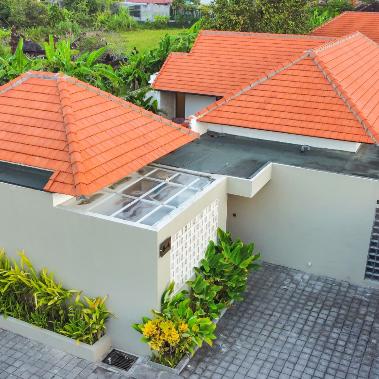 Aerial view of a modern house with orange terracotta roof tiles and a gray paved courtyard surrounded by tropical plants.