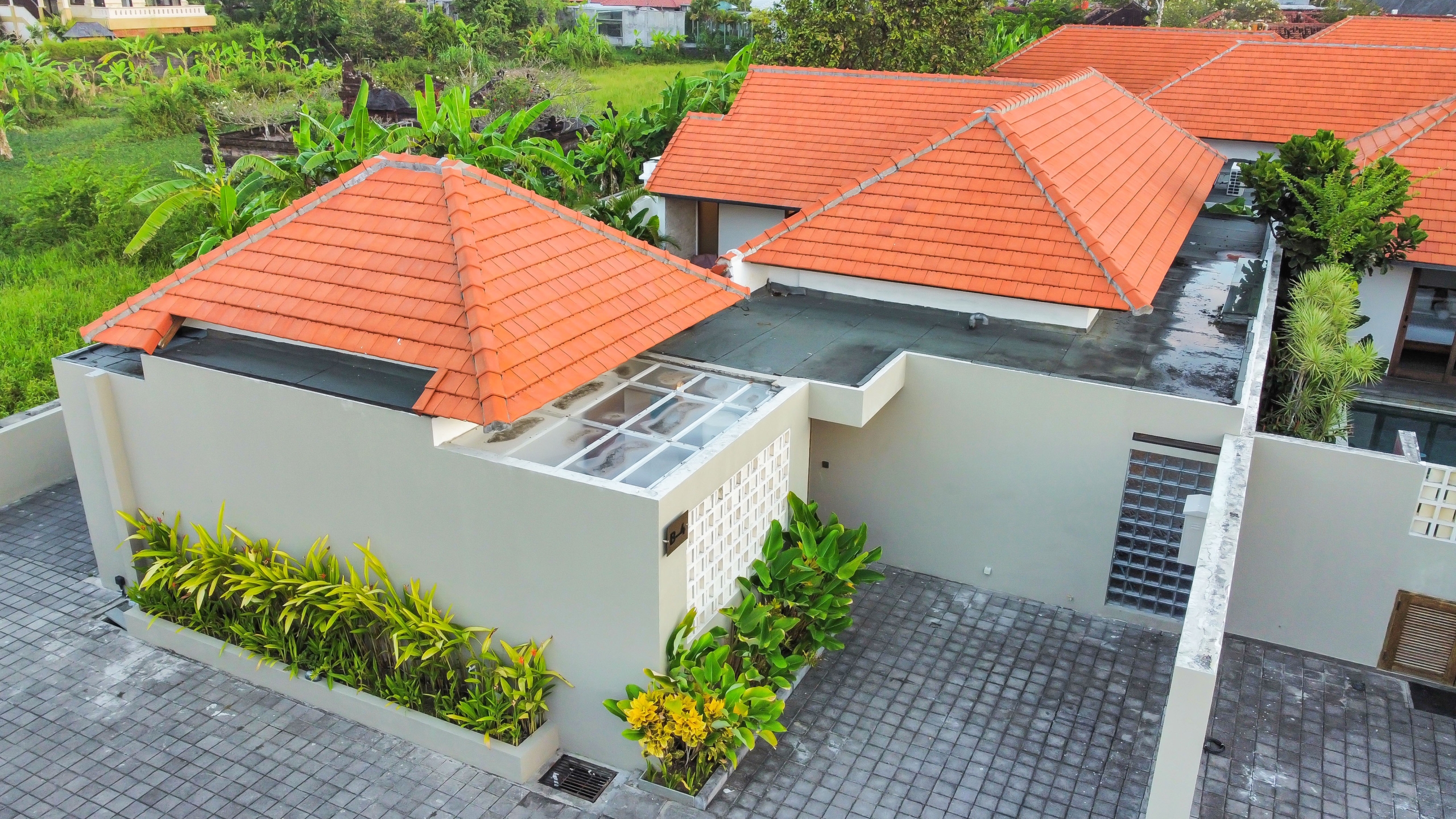dji_fly_20260526_174912_861_1779788958682_photo_optimized Aerial view of a modern house with orange terracotta roof tiles and a gray paved courtyard surrounded by tropical plants.