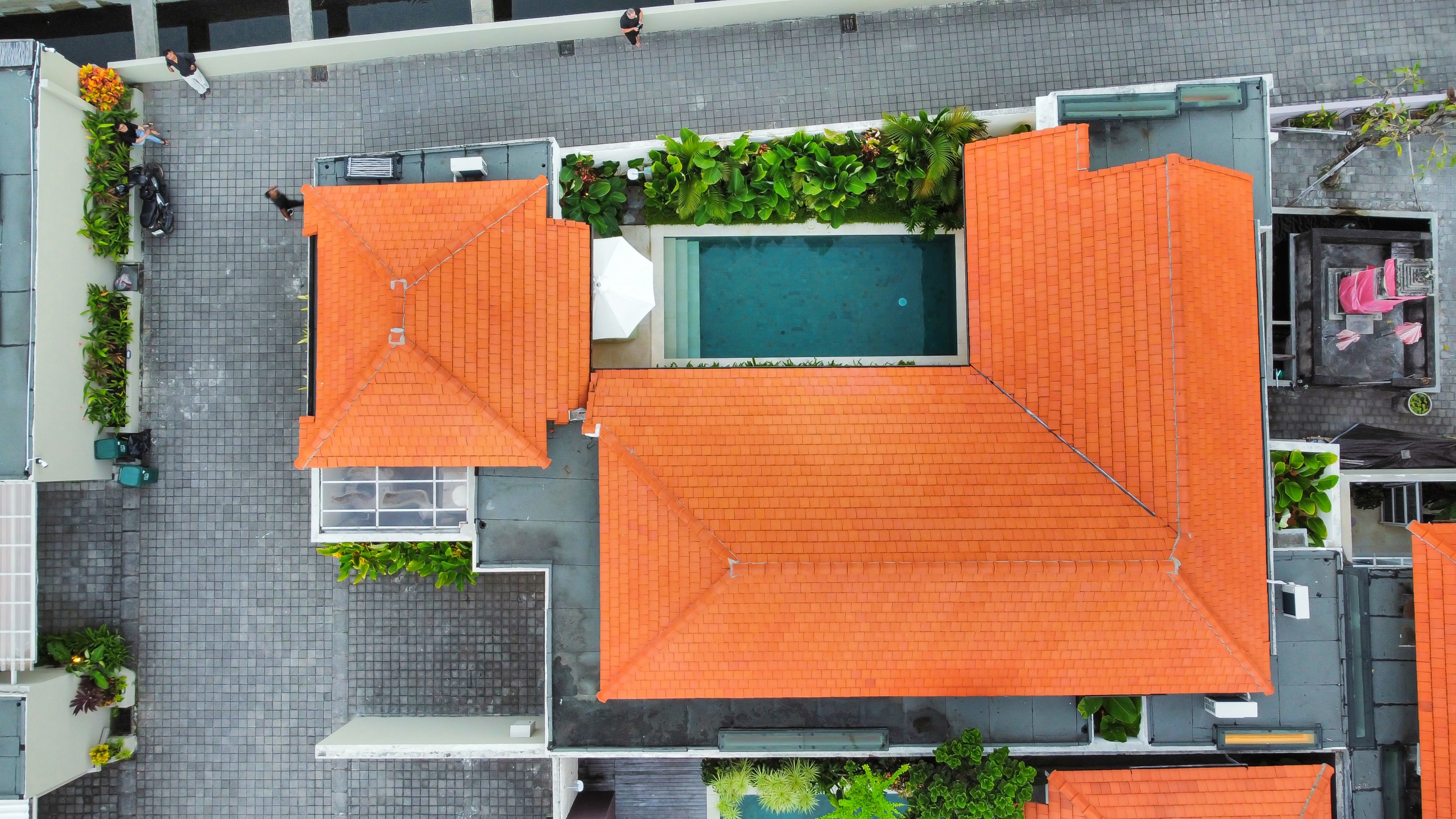 dji_fly_20260526_175928_882_1779789574170_photo_optimized Aerial view of orange-tiled rooftops surrounding a rectangular pool with greenery along the edges and gray tiled walkways nearby.