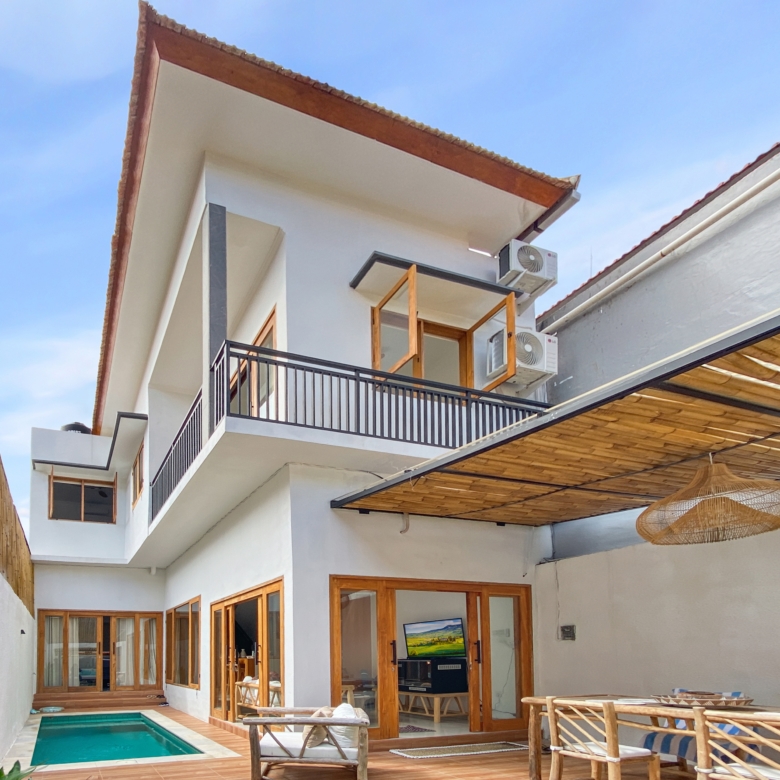 Modern white house with wooden-framed glass doors, a narrow pool, and a shaded outdoor seating area beneath a blue sky.