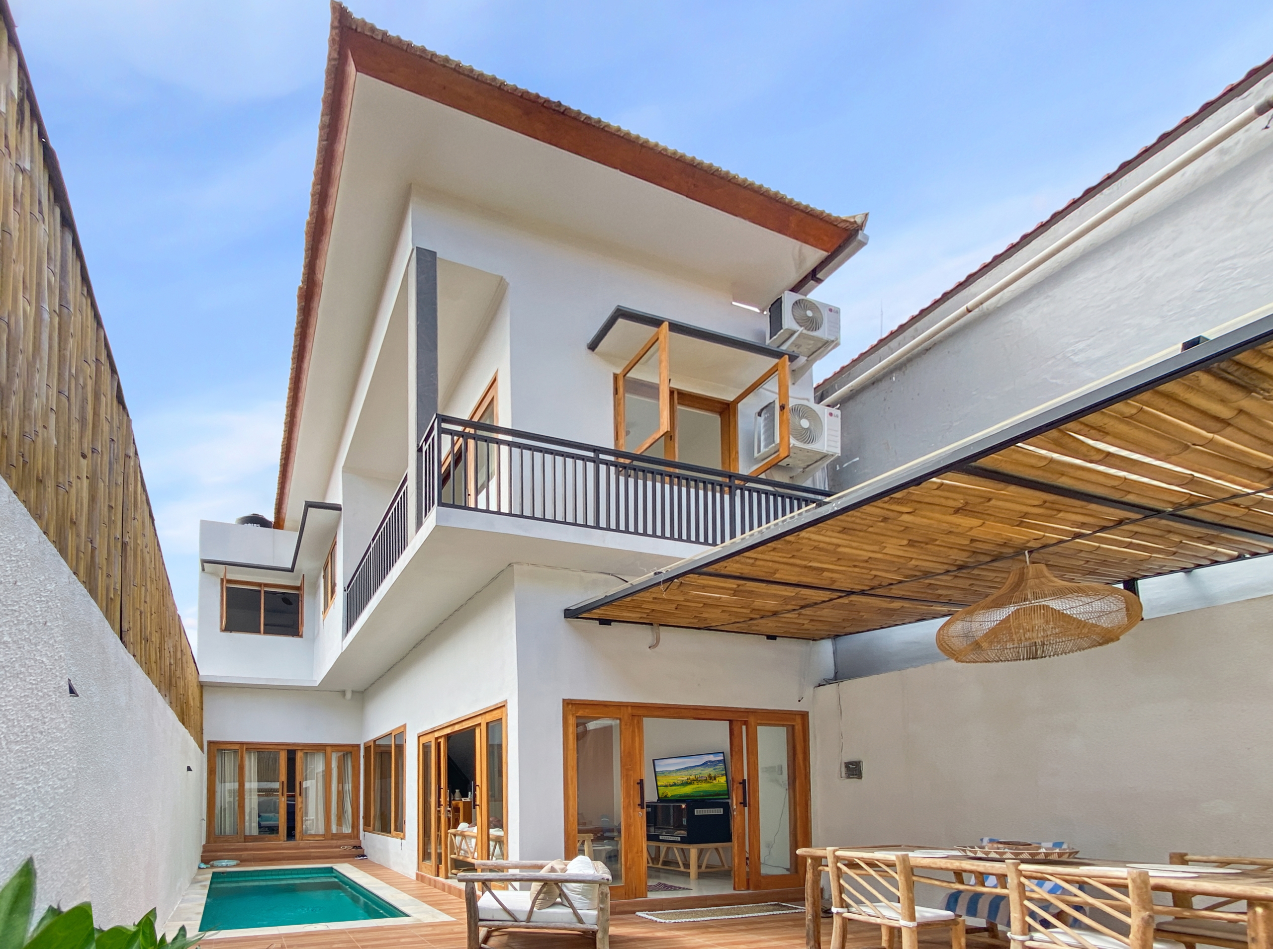 e25bbb67c2a13f8205eaaa31945e4d720b92e0cb Modern white house with wooden-framed glass doors, a narrow pool, and a shaded outdoor seating area beneath a blue sky.
