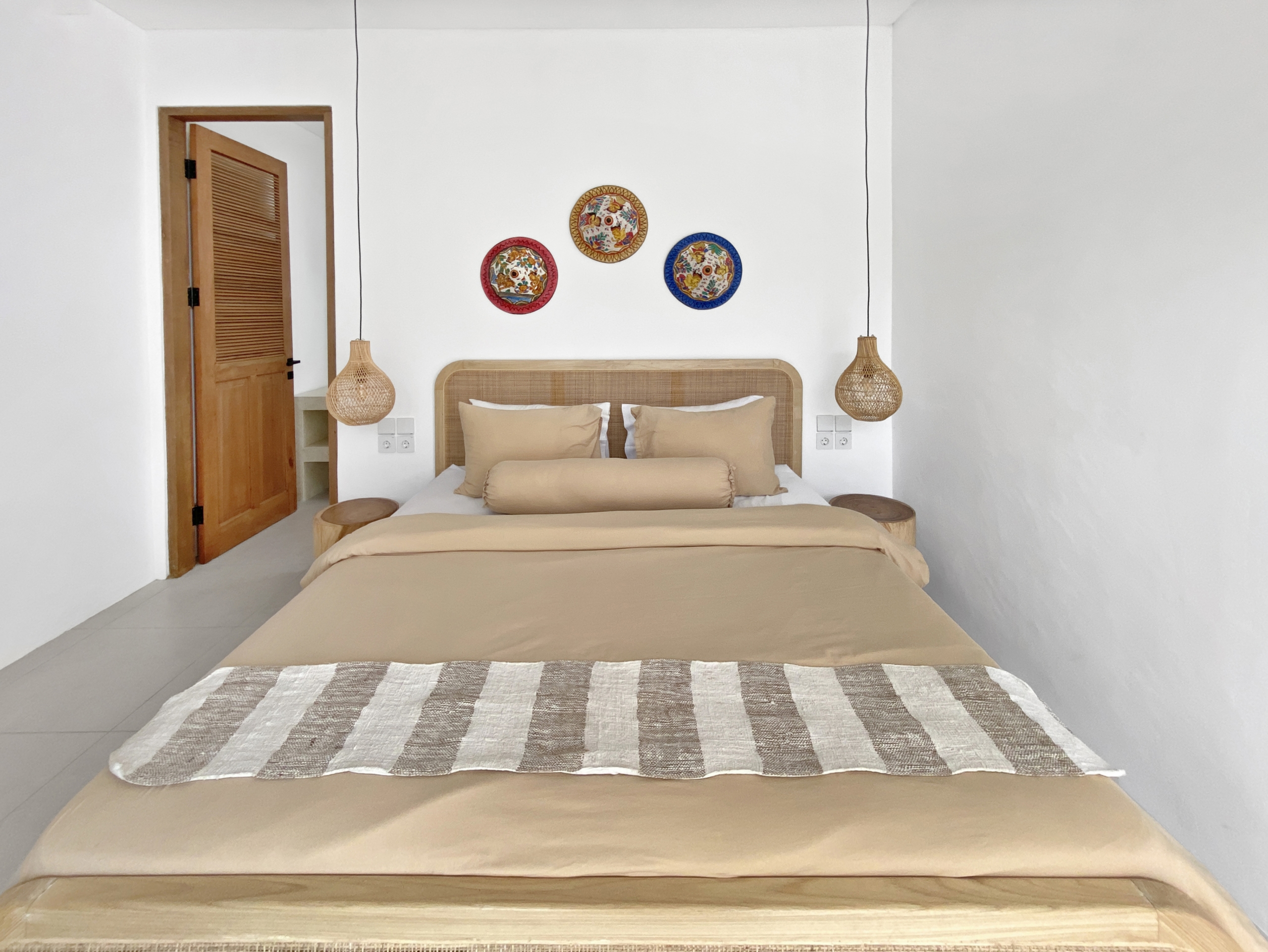e27fb0bccb53efe4e0c64874cd2dc00360976a50_2295-68d5cfaf9fc42 Beige bedroom with a striped bed runner, beige pillows, and a rattan headboard under three colorful circular wall plates.