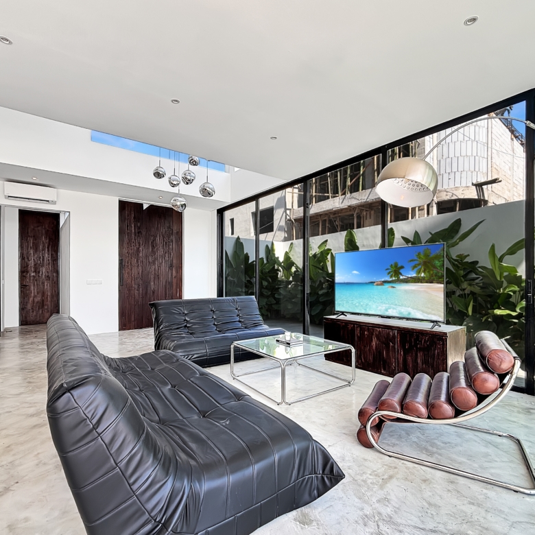 Modern living room with black tufted leather lounge chairs, glass coffee table, and a TV showing a tropical beach scene.
