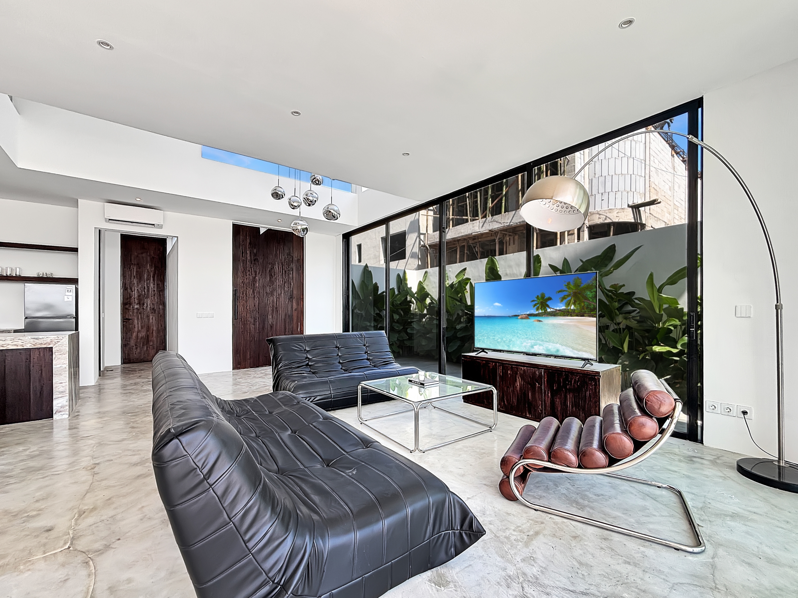 e38f5914ddf4ff76b10eeabcf3b1d4a69a4e8a3f_6810-6a1d7bae0e124 Modern living room with black tufted leather lounge chairs, glass coffee table, and a TV showing a tropical beach scene.