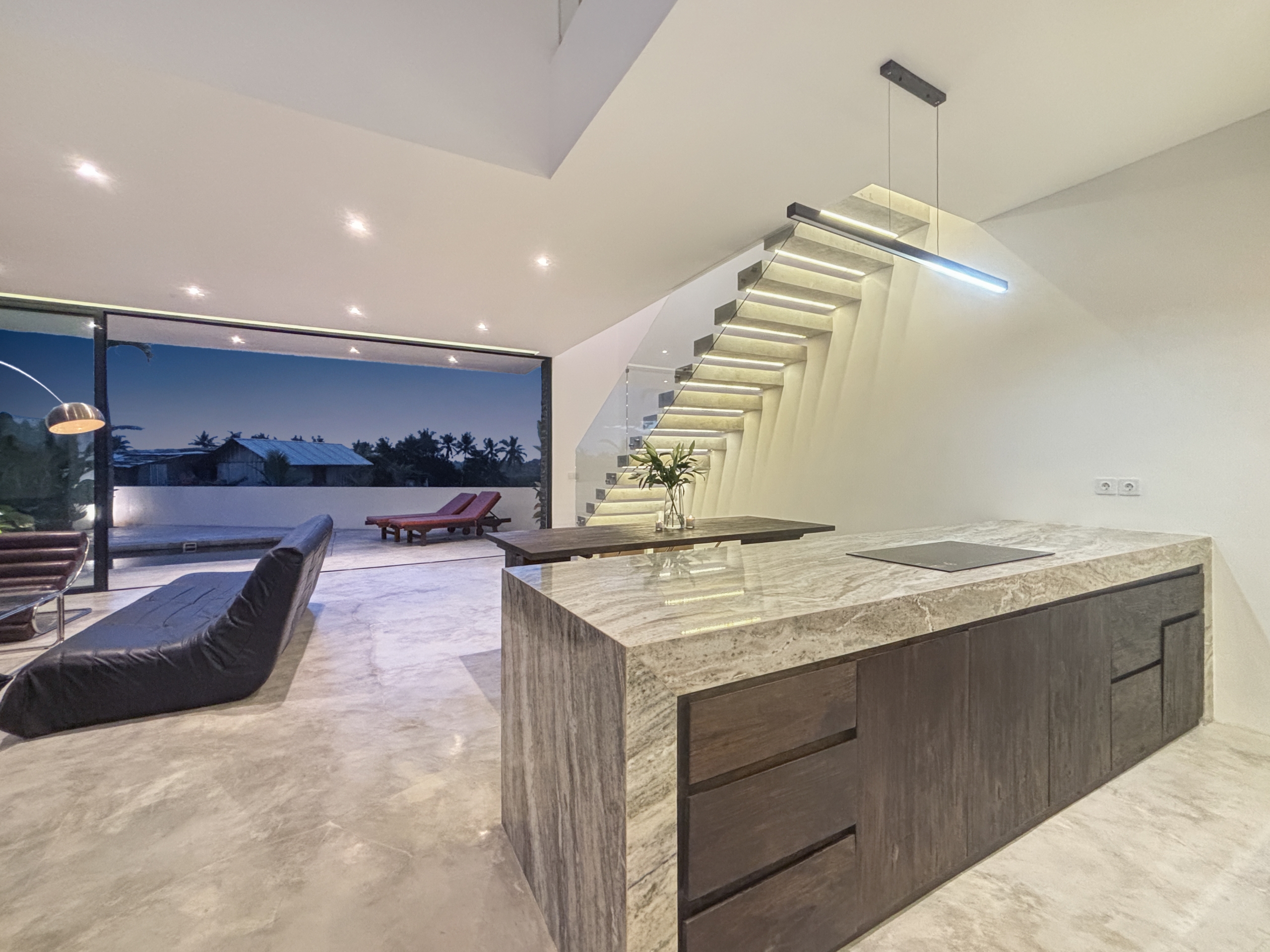 e669cf3878dd86ba5e7175649b4e880632fee8ed_1002-6a05be0662d51 Open-plan interior with a marble kitchen island, dark wood cabinetry, and a floating glass-railed staircase along the wall.