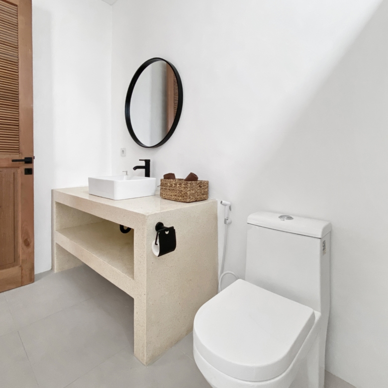 Bathroom vanity with square white sink on beige countertop, round black-framed mirror, wicker basket, and toilet.