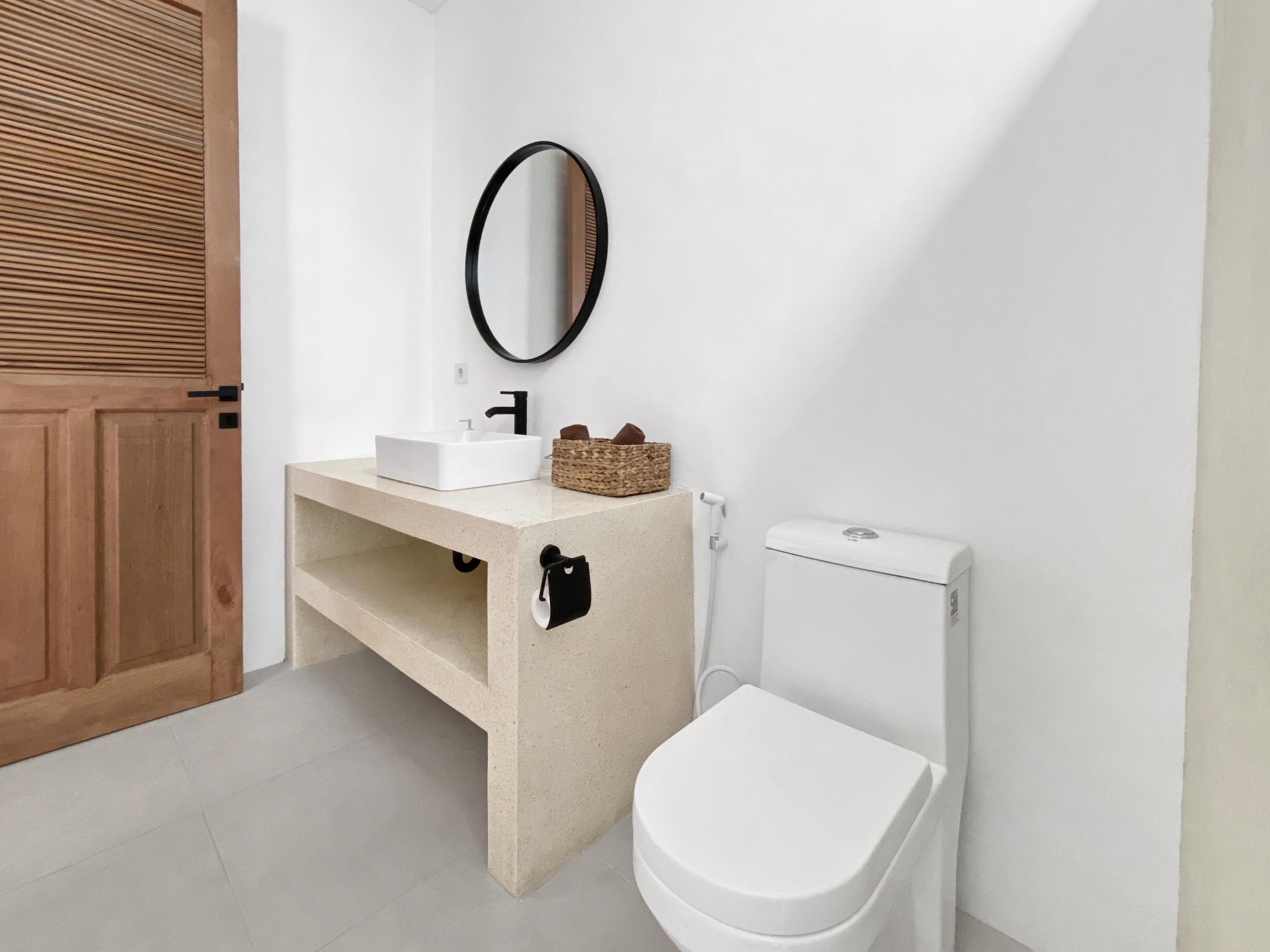 e7c27f3781f13891797c5601161da30799e088f2_2291-68d5cfaef3ec4 Bathroom vanity with square white sink on beige countertop, round black-framed mirror, wicker basket, and toilet.