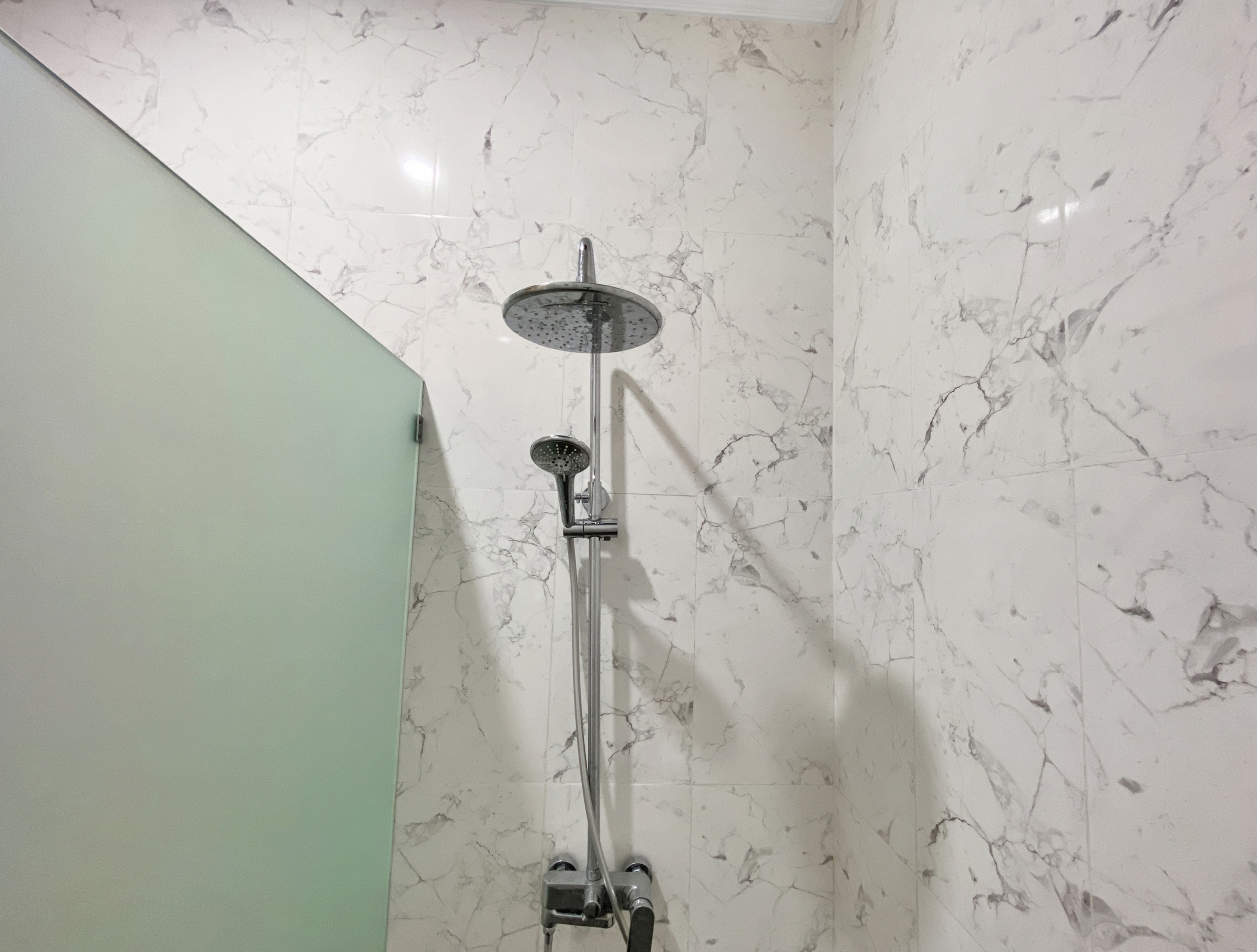eace99861cbd40422112ae70fccb119361abd25c Modern bathroom shower with a large round rain head and handheld wand on a marble-tiled wall enclosure, chrome fixtures visible.