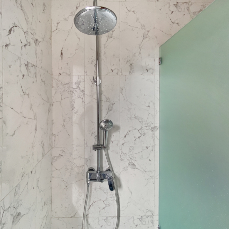Shower setup with a round rainfall showerhead and a handheld wand on a chrome bar against white marble tile, next to a green glass partition.