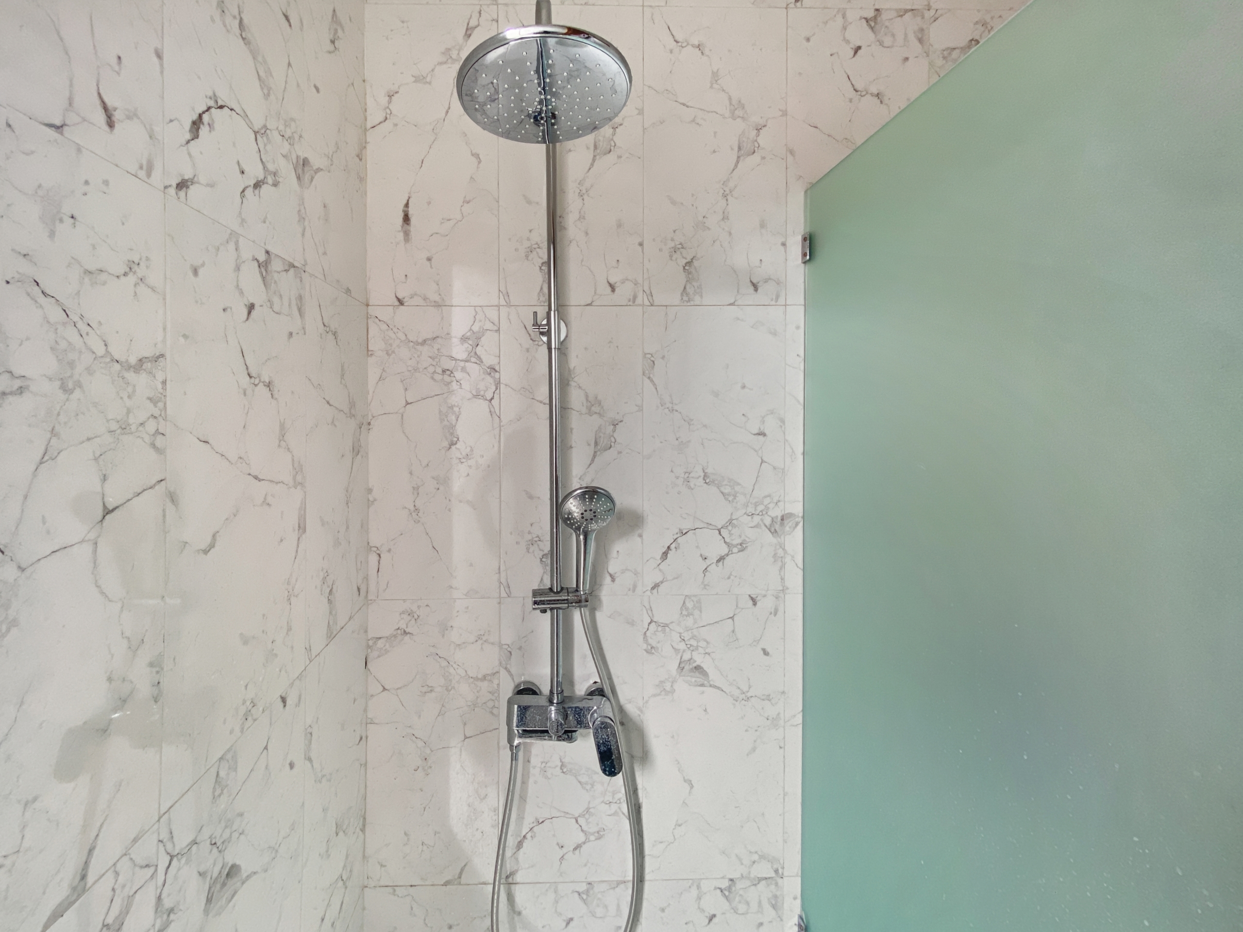 ed5429fdc6ab33f4cb2ad637cebc67da24cabdc3 Shower setup with a round rainfall showerhead and a handheld wand on a chrome bar against white marble tile, next to a green glass partition.