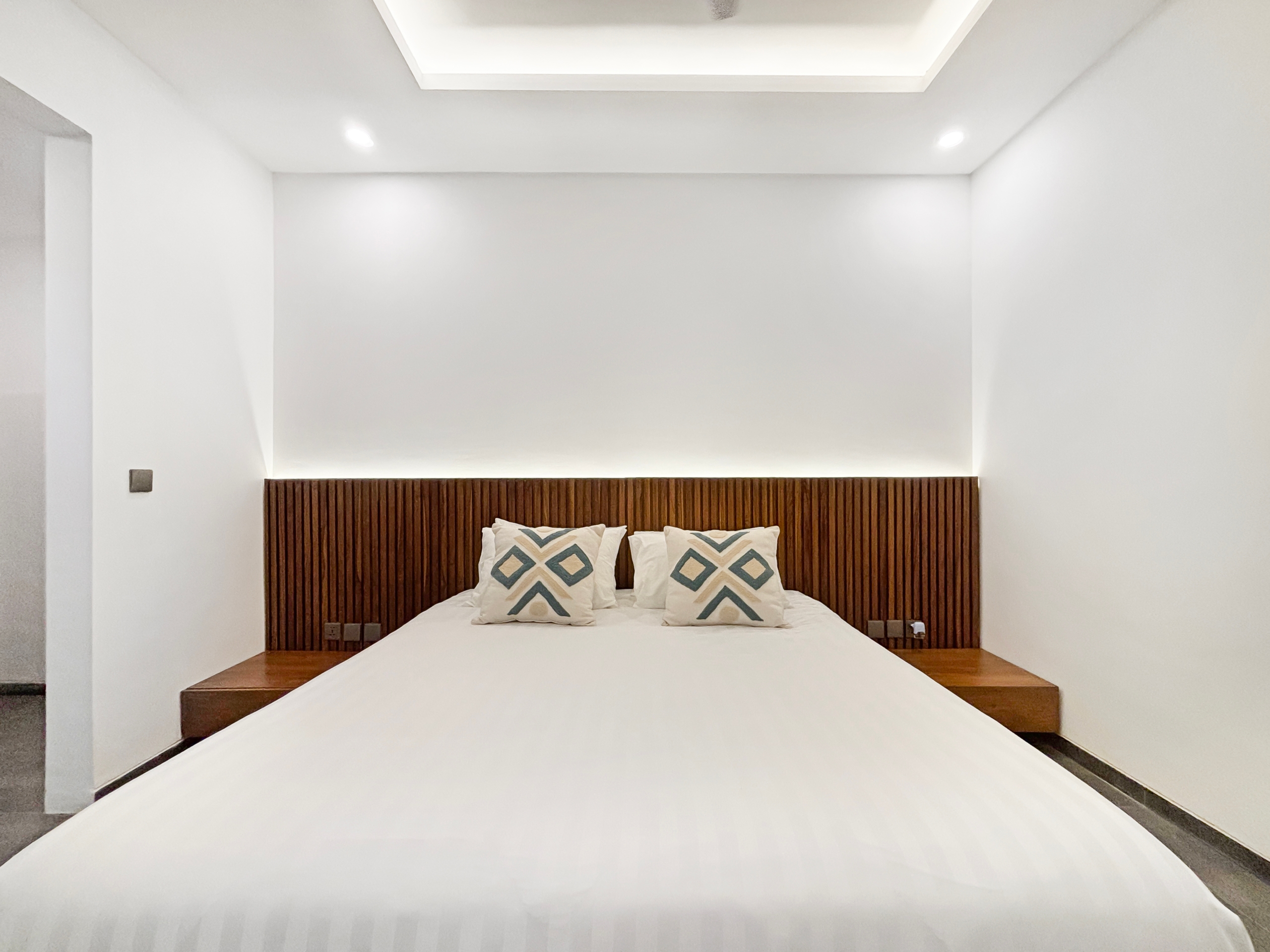 ef0399476d89e2e3232c540401b61050804a6ebe Minimalist bedroom with white bedding and a dark wooden, vertically slatted headboard behind the bed.